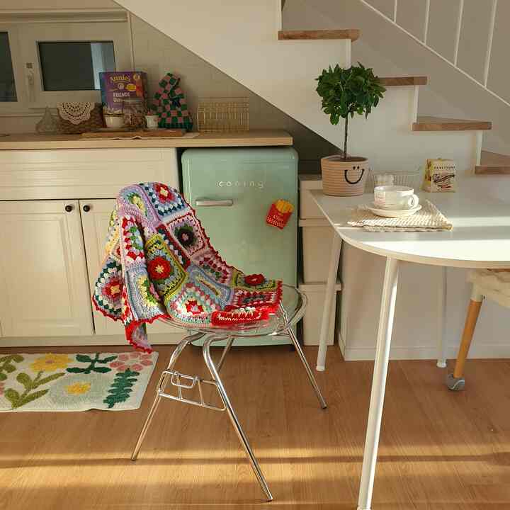 白と木目調が調和したキッチン空間、ミニ冷蔵庫とカラフルなクロッシェブランケットが掛けられた椅子、かわいい小物と植物が配された雰囲気のあるインテリア