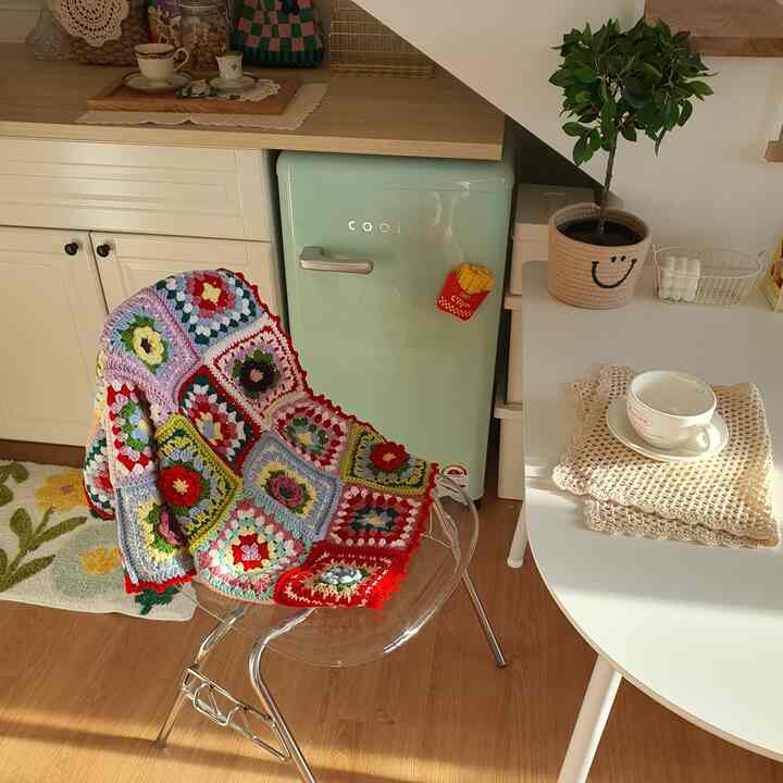 ミントグリーンの冷蔵庫と白いキッチン家具、透明な椅子に多彩な手編みのブランケットが掛けられた温かみのあるミニキッチン