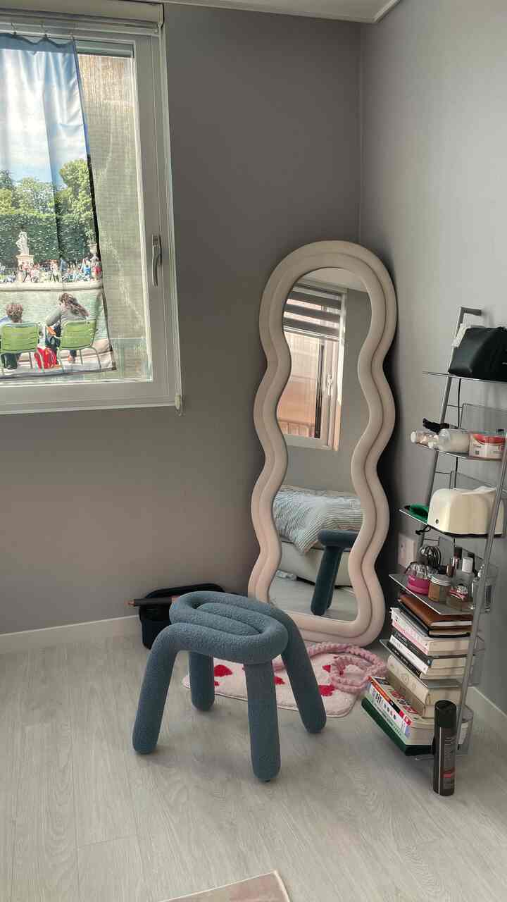 ベージュの壁とグレーの床が調和する小さな部屋、曲線的な全身鏡とブルースツール、金属製の棚が配置されたモダンな空間