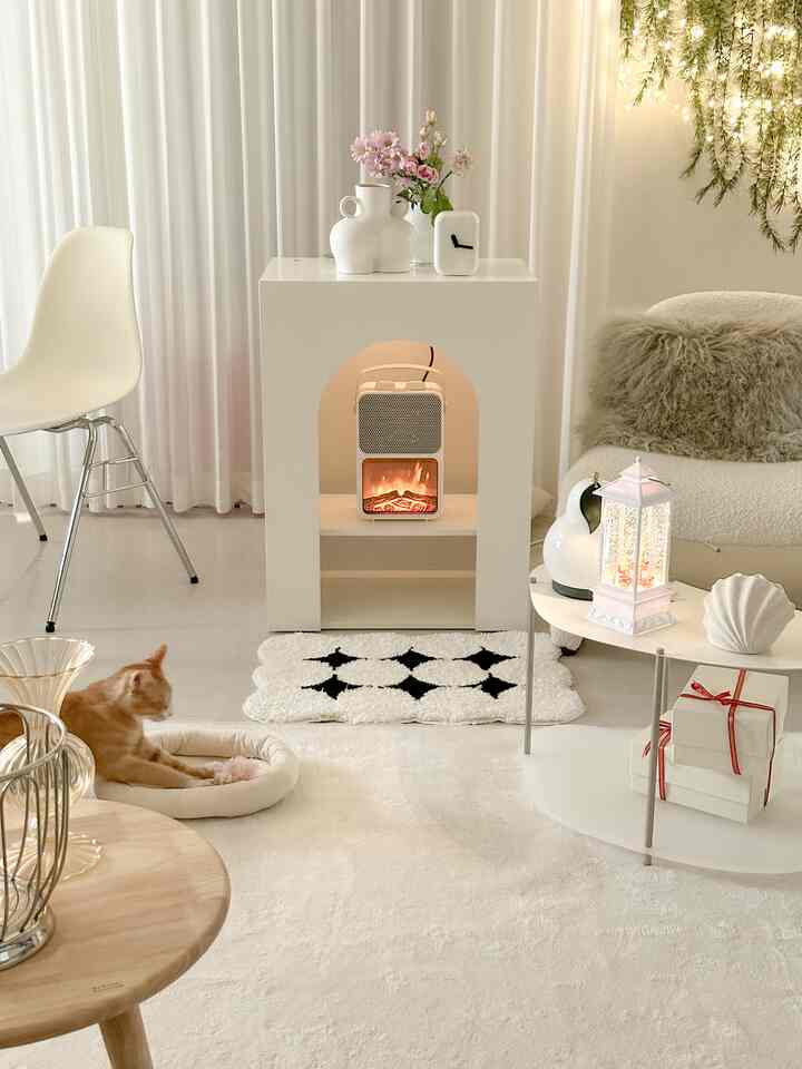Bright ivory-toned living room featuring a central electric fireplace, white sofa, vases with flowers, and a cat resting on the floor, creating a cozy atmosphere