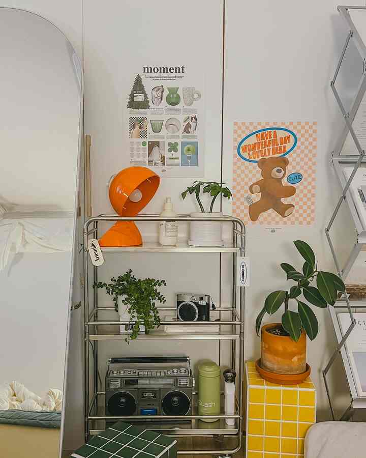 鮮やかなオレンジのアルテミデ照明と植物が配されたモダンでミッドセンチュリー風の寝室、メタルトロリーと全身鏡を含む