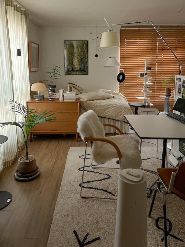 Warm brown wood tones and natural beige blend in this studio bedroom, featuring eco-friendly wood blinds and plants creating a cozy interior