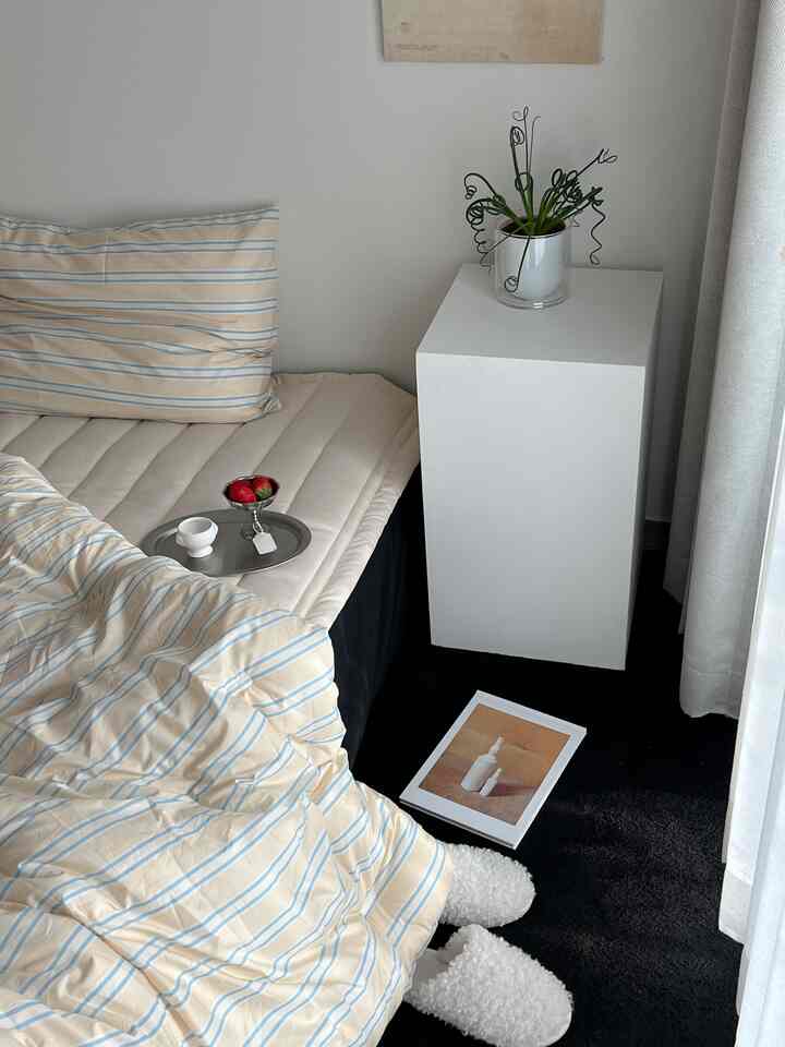 ベージュとブルーのストライプ寝具を配したシンプルモダンな寝室、白いナイトテーブルと黒いカーペットが調和する居心地の良い空間