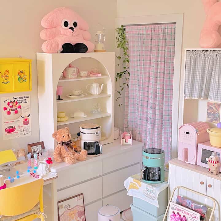 パステル調のトーンとピンク色が際立つワンルーム、かわいいぬいぐるみと多彩なキッチン小物が配置された雰囲気のあるインテリア