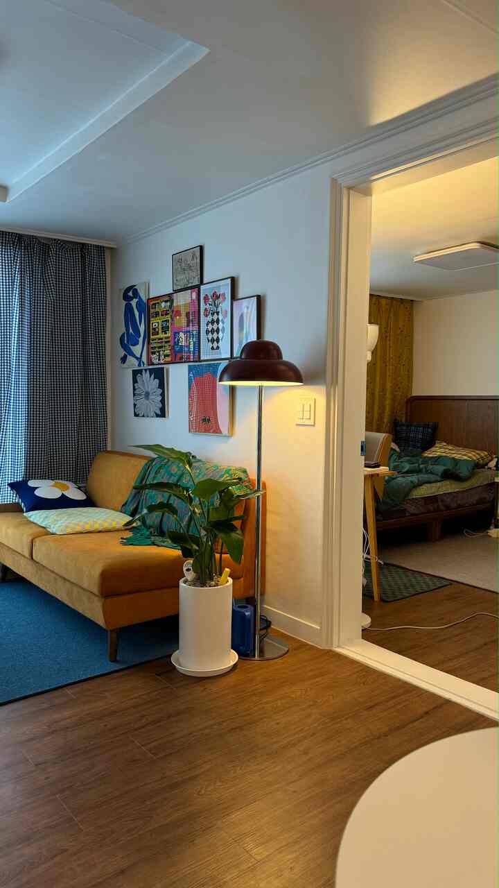 マスタード色のソファとブルーのカーペットがあるナチュラルモダンなリビング、壁に多様なアートが飾られ、寝室が見える温かみのある空間