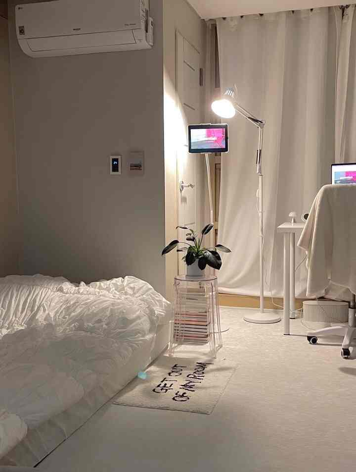 ホワイトトーンのミニマルな寝室、シングルベッドとプランツを置いた透明なスツール、ホームオフィスとして機能するデスクとフロアランプを配した雰囲気のあるインテリア