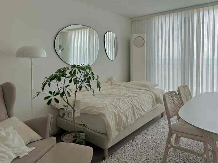 ホワイトとベージュのトーンのワンルームリビングベッドルーム、大きな鏡と快適なアームチェア、ダイニングテーブルが配置されたナチュラルモダンな雰囲気のあるインテリア