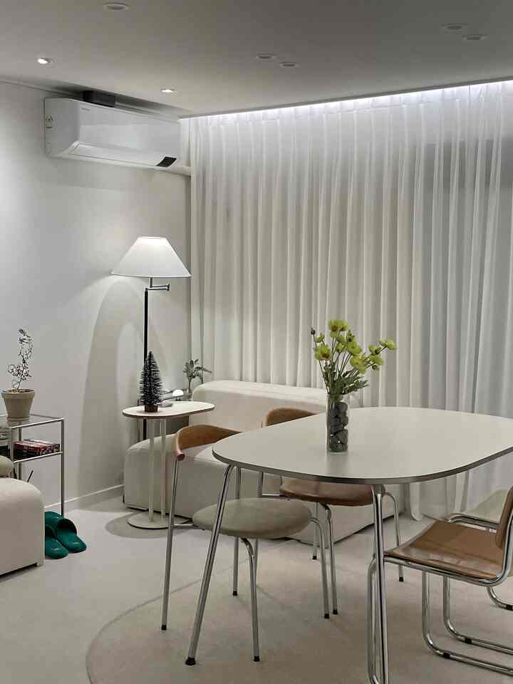 白とベージュの色調のリビング兼ダイニングスペース、シンプルモダンなダイニングテーブルと椅子、フロアランプ、窓全体を覆う白いカーテンが配置された印象的な空間