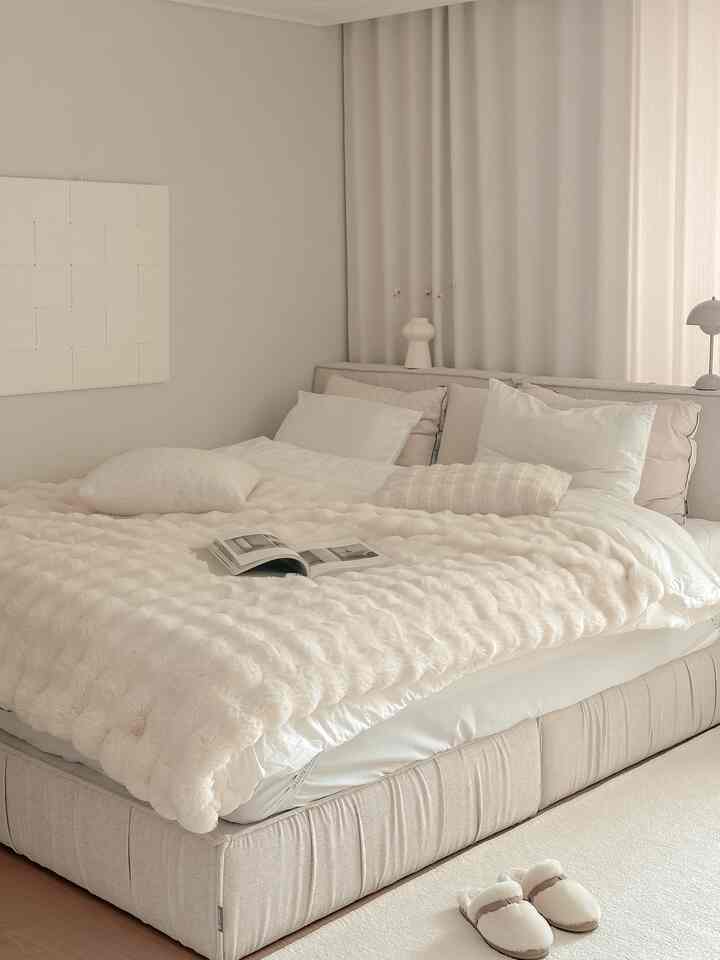 明るいベージュと白の色調の温かみのある寝室、大きなベッドとふわふわのブランケット、クッションが整然と配置された空間