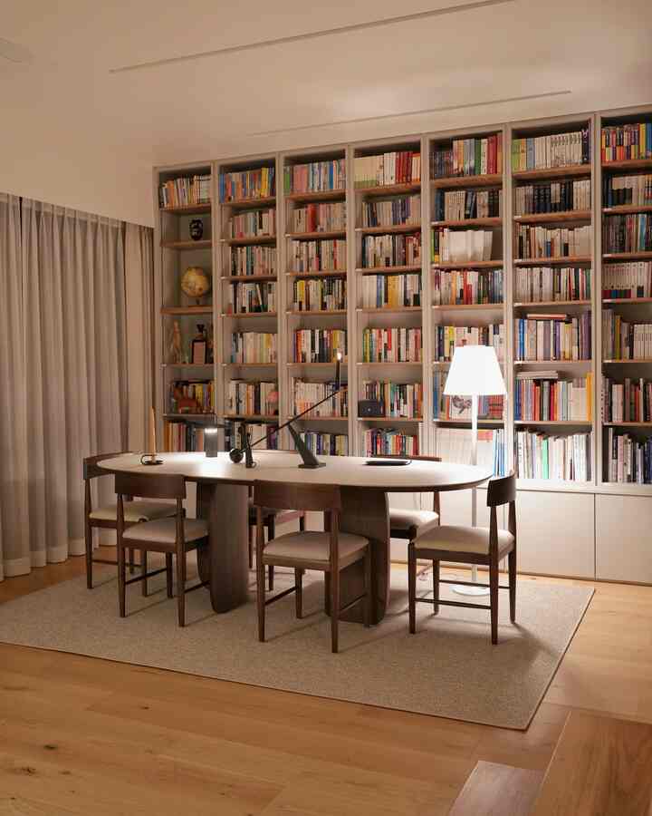 White and wood-toned study-style home office featuring a large bookshelf, oval dining table, and warm lighting ambiance