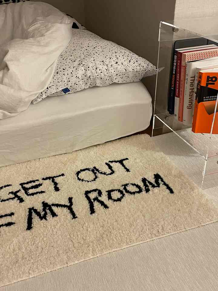 ホワイトとブラックの色調の6畳寝室、白いマットレスと斑点模様の枕、‘GET OUT OF MY ROOM’と書かれたラグが置かれたミニマルなインテリア