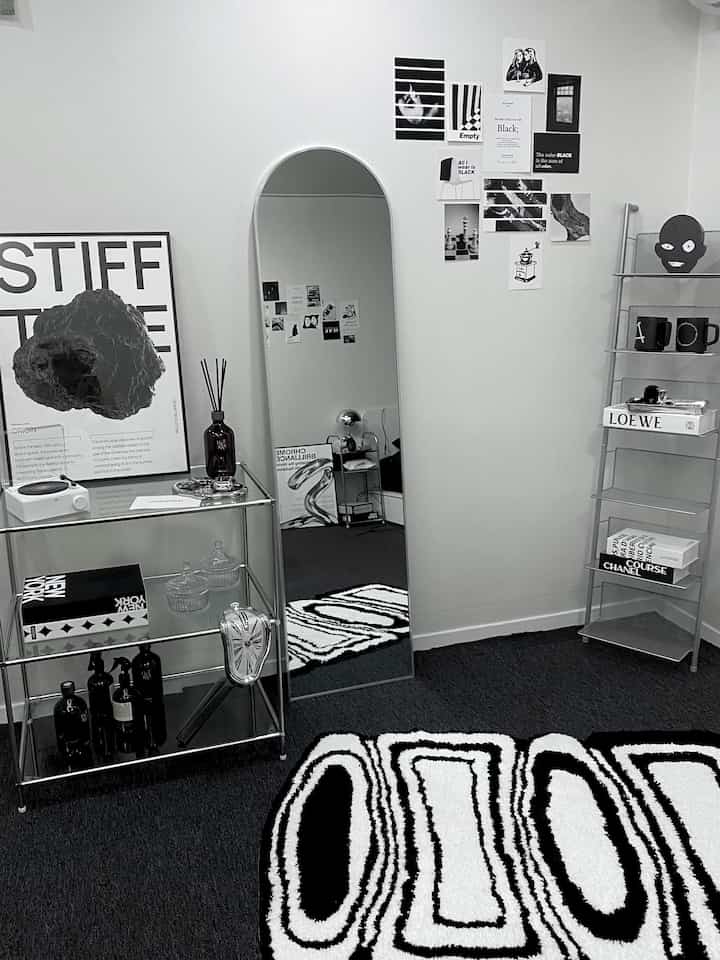 Black and white toned studio living room with steel shelves, a large arch mirror, and patterned rug in a modern minimalist style