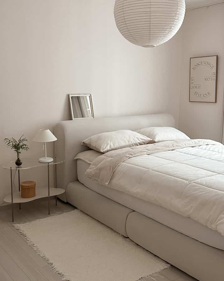 アイボリーのミニマルな寝室、丸みのあるファブリックベッドフレームと透明なサイドテーブルが配置された落ち着いた空間