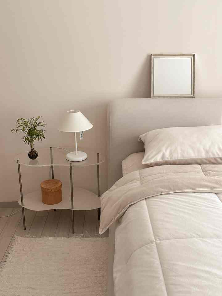 柔らかなベージュ色調の寝室、ファブリックベッドフレームと透明なナイトテーブル、シンプルな照明が調和した落ち着いた空間