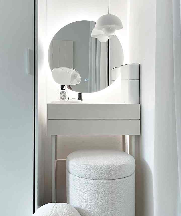 White-toned bedroom vanity area featuring a large circular mirror, pendant lights, and a soft-textured vanity stool in a simple interior