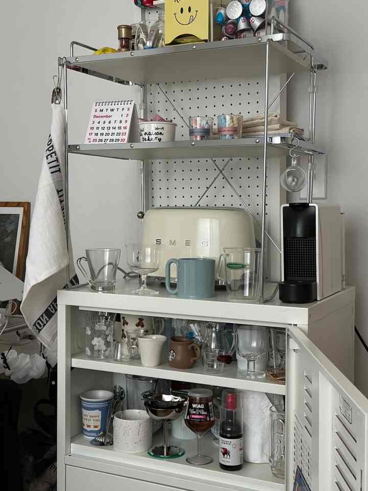 白い壁と収納キャビネットのあるキッチンスペース、SMEGのトースターとコーヒーマシン、多彩なカップやグラスが整頓された雰囲気のあるホームカフェインテリア