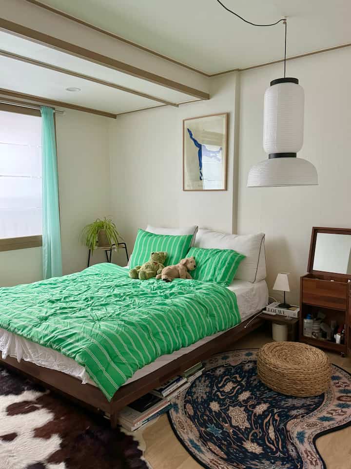 A bedroom with white walls and natural wood tones, featuring green bedding and a patterned rug creating a cozy atmosphere