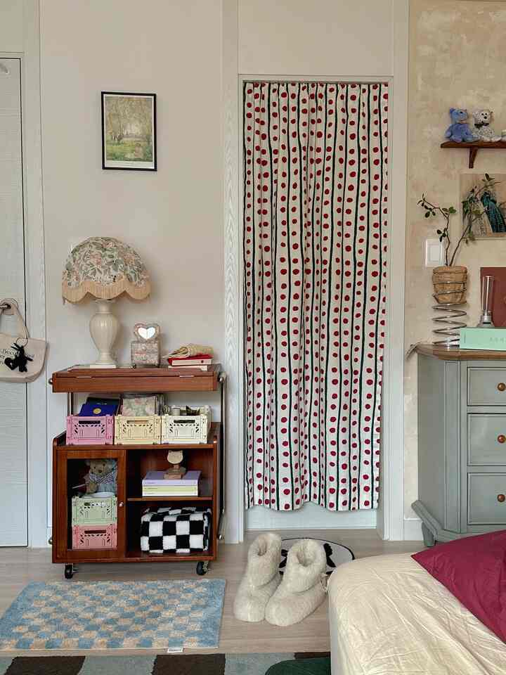 White and beige toned bedroom featuring vintage lamp, dresser, and a red polka dot curtain on the door creating cozy ambiance