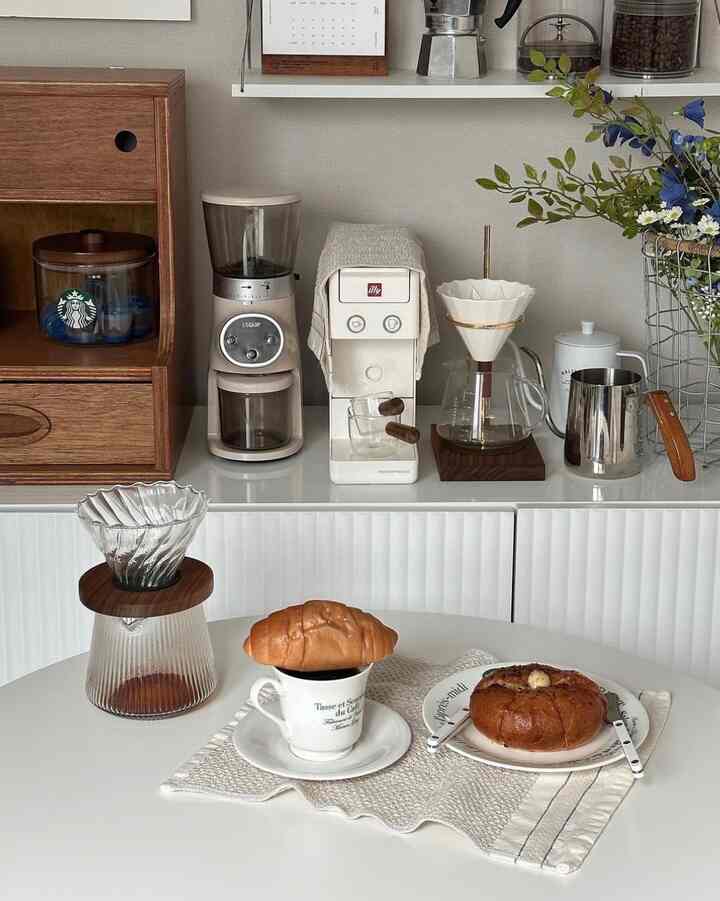 White and brown toned home cafe space featuring a coffee machine, drip coffee set on a clean dining table with bread, creating cozy atmosphere