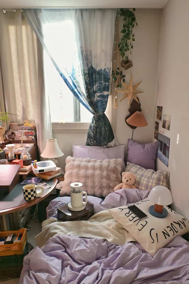 ラベンダー色の寝具とブルーのカーテンがある小さなワンルーム寝室、ヴィンテージ雑貨と居心地の良い空間