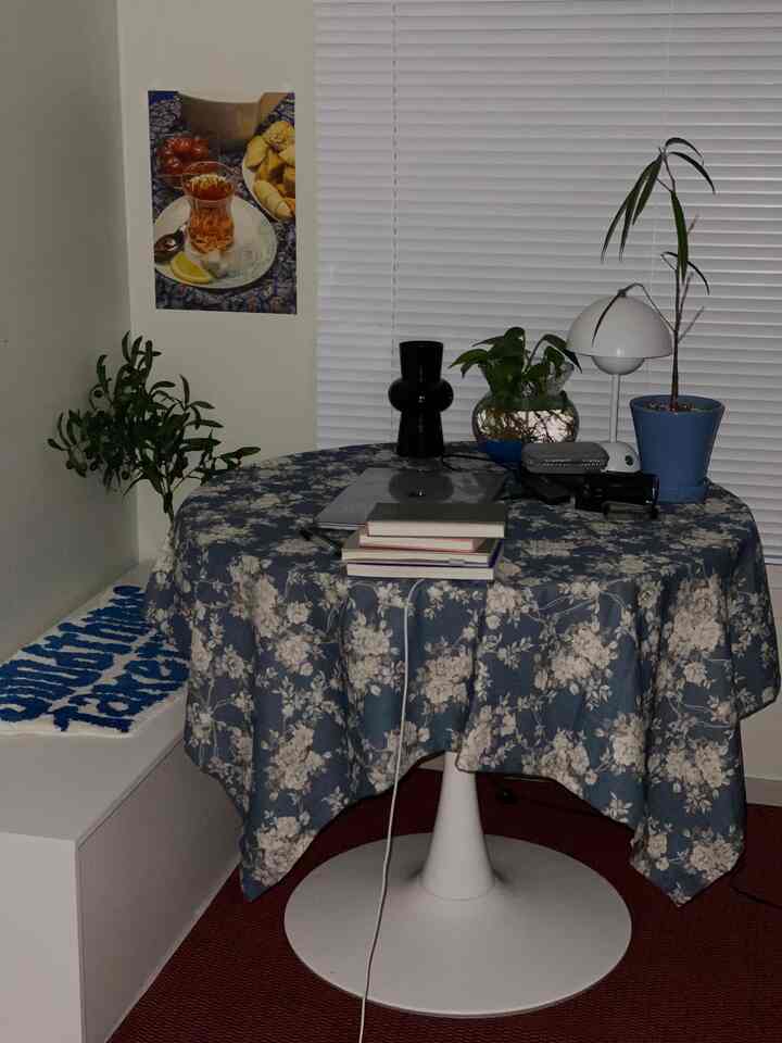 Retro-inspired space with a round table covered by a floral tablecloth, plants, books, and lighting in front of white blinds