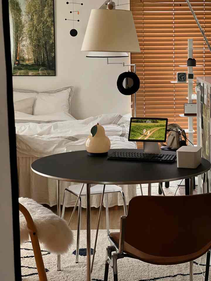 A minimalistic studio apartment bedroom and home office with white and wood tones, featuring simple furniture and wooden blinds