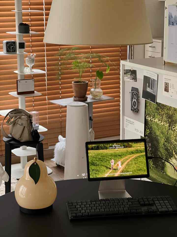 Minimal home office featuring a modern black desk in front of wood tone blinds background, complemented by a cream table lamp and green plants