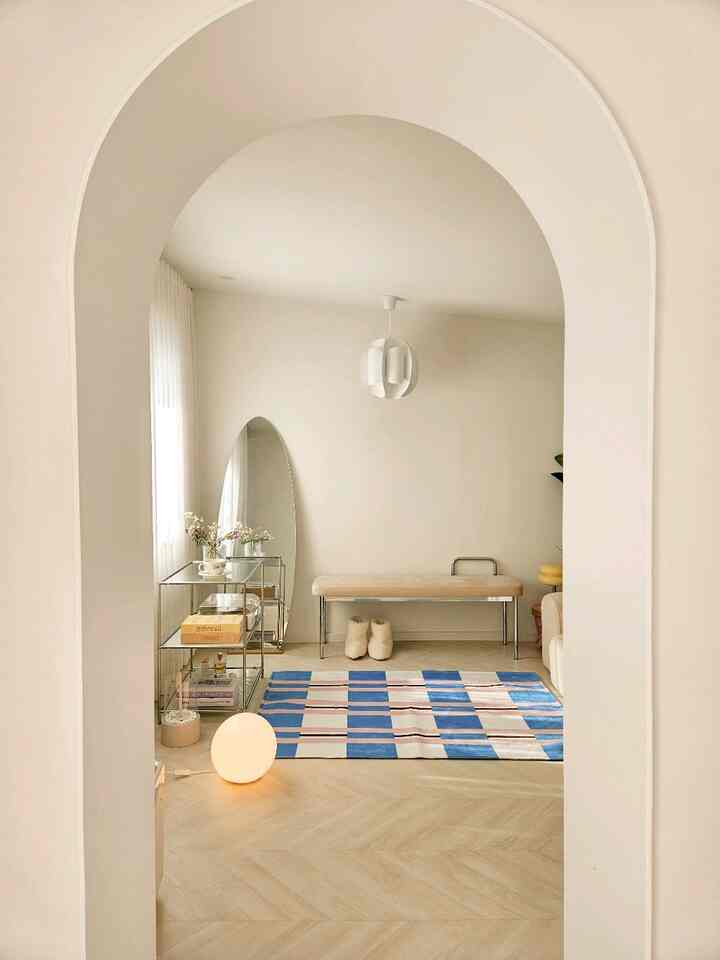 柔らかなホワイトと木目調のロフト空間、アーチ形の入り口とベンチ、鮮やかな青のパターンラグが映えるモダンでミニマルなインテリア