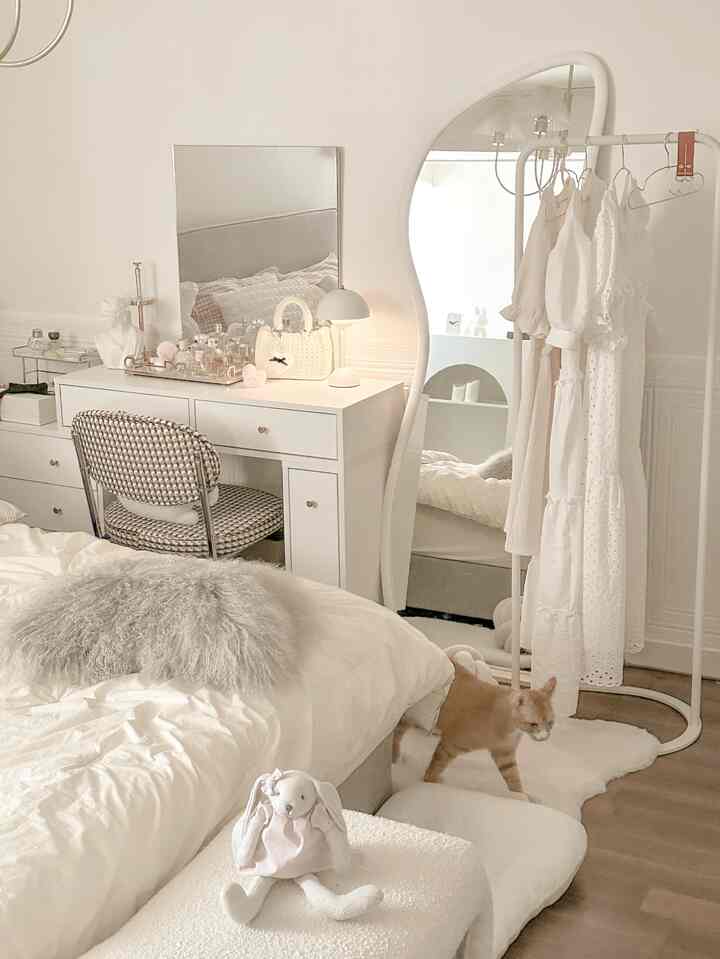 Ivory natural-toned bedroom featuring white vanity, large freeform mirror, and clothes rack with dresses in a neat setting
