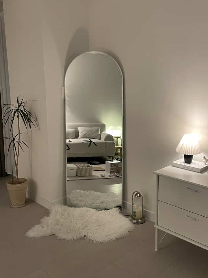 Living room with white walls and furniture, featuring an arched full-length mirror and white fake fur rug creating a cozy modern atmosphere.
