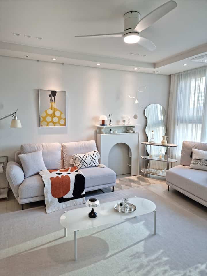 Soft white-tone living room featuring fabric modular sofas centered with a long white table, modern ceiling fan and light curtains creating a cozy atmosphere