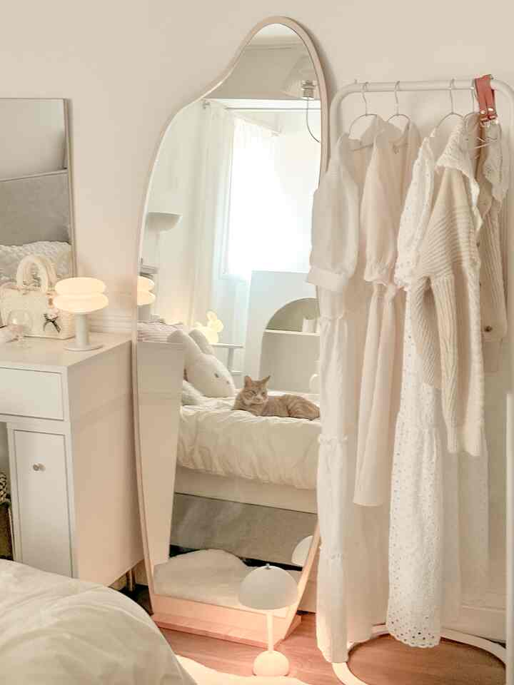 ホワイトとナチュラルカラーの寝室、不定形の全身鏡、猫と服が掛かったハンガーが配置された居心地の良い空間