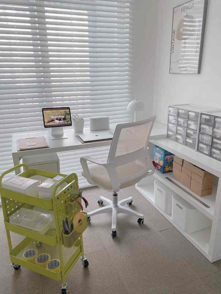 A minimal workspace in white and natural tones, featuring a clean white desk, organized storage trolley, and window blinds creating a bright atmosphere