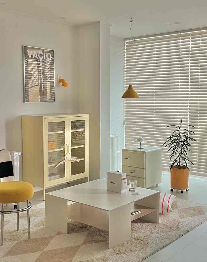 Bright and clean living room with white and yellow tones, featuring a white coffee table center, yellow chair, storage cabinets, and a plant by the window