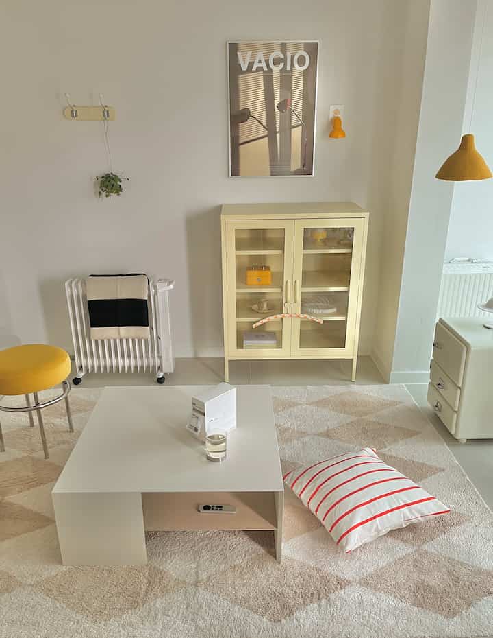 Natural tone living room featuring a central low coffee table, yellow stool, and cabinet with radiator on wall in a simple layout