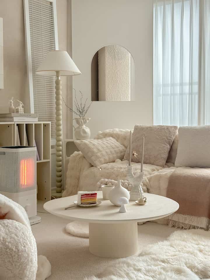 A cozy living room in soft beige and white tones featuring plush cushions, a round coffee table, and a floor lamp