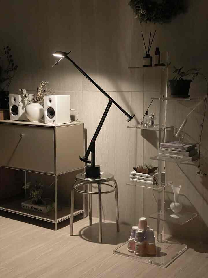 Living room in white and black tones featuring a modern floor lamp, stainless steel stool, and transparent shelving with a natural atmosphere