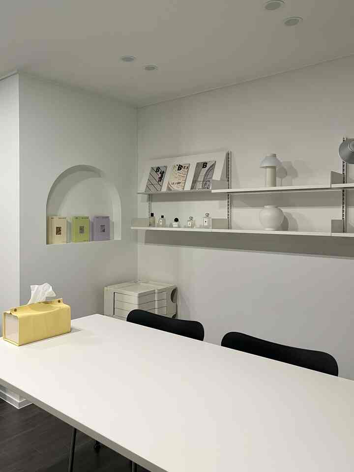 Bright white-toned study-like space featuring a central white table with black chairs, wall shelves, and an arched niche displaying magazines and decor in a minimal style