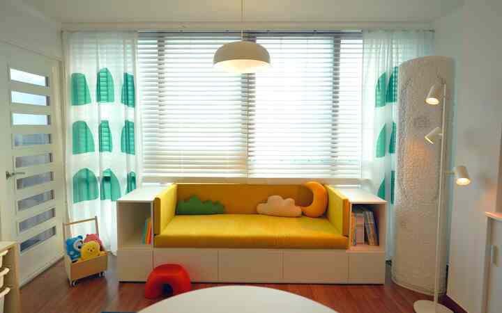 白と黄色の色調が映えるリビングの窓辺、収納ソファとグラフィックシフォンカーテン、子供用クッションとおもちゃが置かれた雰囲気あるインテリア