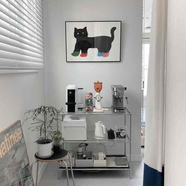 White-toned small kitchen corner featuring metal shelving with coffee machines, blinds, and wall art creating a clean, minimal home cafe atmosphere