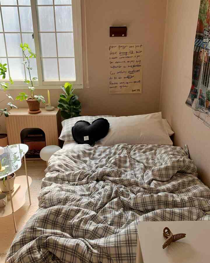 柔らかなベージュ色の壁とチェック柄の掛け布団カバーがある居心地の良い寝室、窓際に複数の植物が配置され自然な雰囲気を醸し出すインテリア