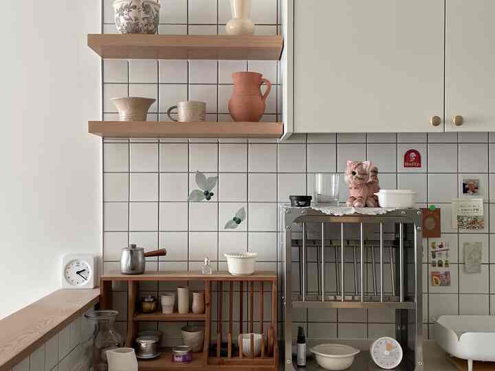 Modern kitchen with white tiled walls and wooden shelves, featuring a metal dish rack and neat decorative items