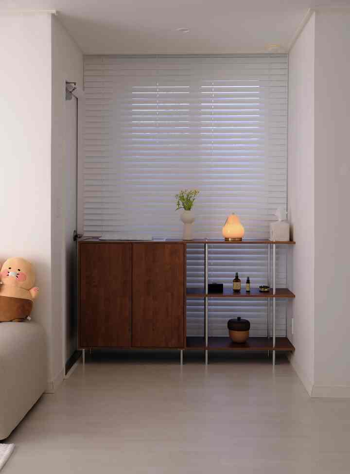 ホワイトトーンのリビングの窓辺にブラウンの3段シェルフと照明、花瓶などが配置された清潔感のある落ち着いた空間