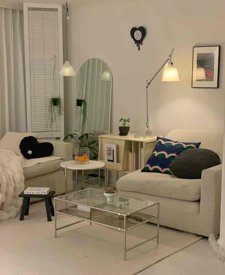 White-toned living room featuring cream sofas with various cushions, an arched full-length mirror, and a heart-shaped wall clock in a modern interior