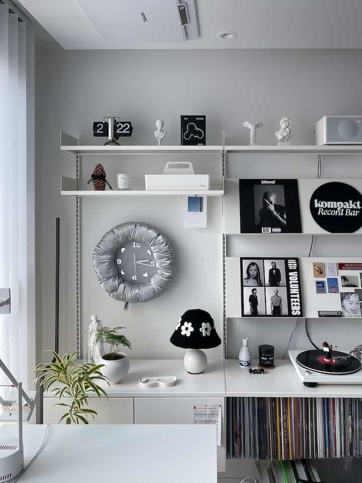 白とグレーの色調のリビングルーム兼ホームオフィス、白い壁と棚、大きなグレーの時計、黒いテーブルランプと植物が配置された印象的なインテリア