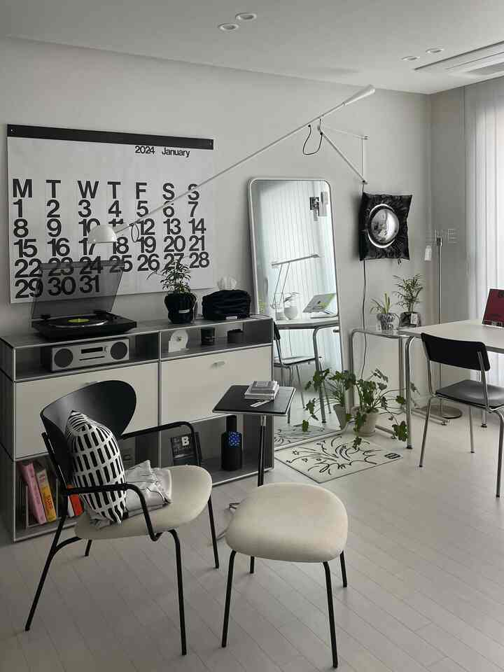 A modern living room and home office space in white and black tones featuring a lounge chair, stool, wall lamp, and a large calendar poster