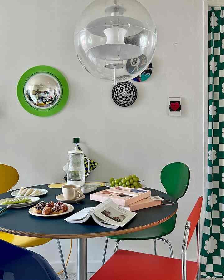 A white-dominated dining room with colorful chairs, featuring a round dining table set with teapot and plates in a clean modern space