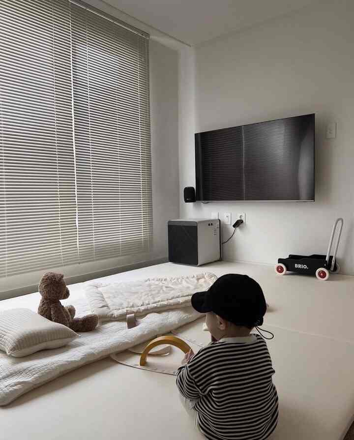 ナチュラルカラーの寝室、前景に赤ちゃんが座り、大きな窓にブラインドがあり、壁に取り付けられたテレビが配置されたミニマルな雰囲気のインテリア