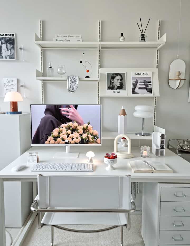 Primarily white-toned home office featuring a white desk, chair, wall shelves, and various decorative objects in a neat workspace