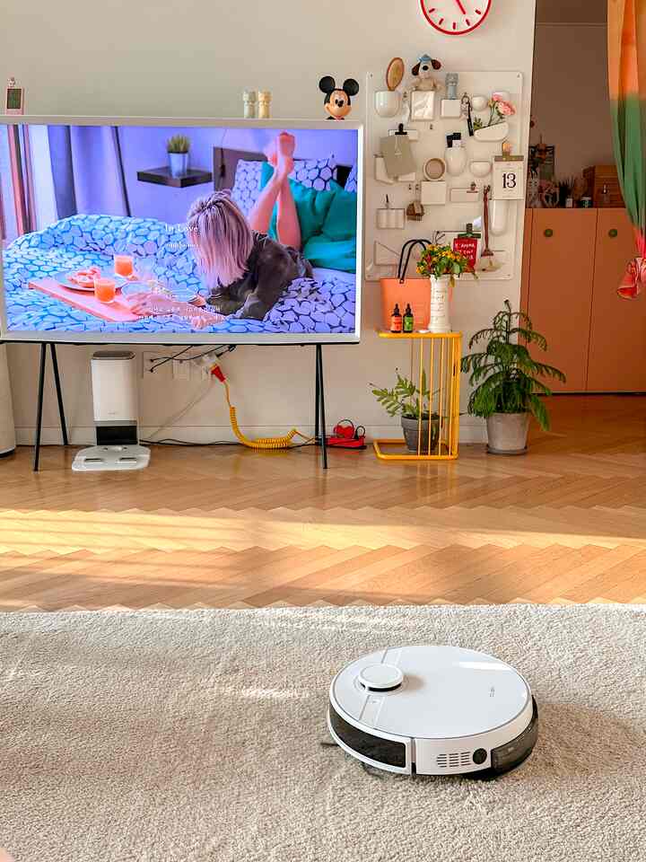 白と木目調が調和したリビング、巨大なテレビとロボット掃除機、植物が配置された明るく清潔な空間
